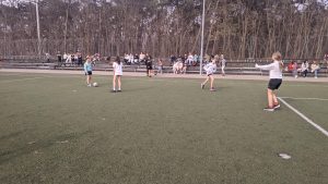 Na zdjęciu widzimy uczenice klas 5 walczące o piłkę w trakcie meczu piłki nożnej. Med odbywa się na boisku ze sztuczną nawierzchnią, zawodniczki ubrane w stroje sportowe, oznaczone są szarfami w kolorach żółtym o czerwonym.