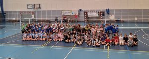 Jest to grupowe zdjęcie wszystkich uczestników turnieju MiniLider CUP. Około 100 zawodników i zawodniczek ubrani w strojach klubowych. W tle widać siatkę do piłki siatkowej oraz biało-szarą ścianę hali. Parkiet jest niebieski z licznymi liniami. Zdjęcie jest bardzo kolorowe.