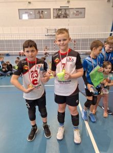 To zdjęcie przedstawia dwóch zawodników UKS Czerwonak, którzy prezentują zdobyty puchar i medale za III miejsce w kat. „Dwójki” chłopców. Są ubrani w stroje meczowe. Koszulka czarno-czerwono-biała a spodenki czarne. W tle widać siatkę do piłki siatkowej. Po obu stronach zdjęcia można dostrzec kilkoro zawodników z innych zespołów. Parkiet jest niebieski z licznymi liniami.