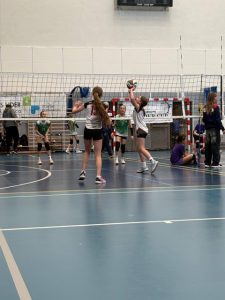 Zdjęcie przedstawia akcję meczową. Dwie zawodniczki UKS Czerwonak rozgrywają piłkę. Jedna jest w pozycji wystawy z piłką na dłoniach a druga szykuje się do ataku z lewego skrzydła. Przed zawodniczkami jest siatka ustawiona na wysokości 200 cm. Za siatką ustawione są przeciwniczki w pozycji obrony. W tle widać ścianę hali na której znajdują się banery. Widać też stolik sędziowski przy którym stoją sędziowie boiskowi. Parkiet jest niebieski z licznymi białymi liniami.