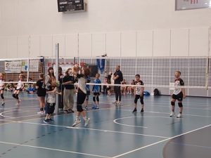 Widać na zdjęciu mecz dwóch drużyn UKS Czerwonak w kat. „dwójki” chłopców. Grają przeciwko sobie. Wszyscy są ubrani w stroje meczowe. Koszulka czarno-czerwono-biała a spodenki czarne. Pomiędzy nimi siatka ustawiona na wysokości 200 cm. W tle widać innych zawodników, którzy czekają na swoje mecz oraz trenera UKS Czerwonak. Tło zdjęcia to biało-szara ścian hali w Rokietnicy. Parkiet jest niebieski z licznymi białymi liniami.