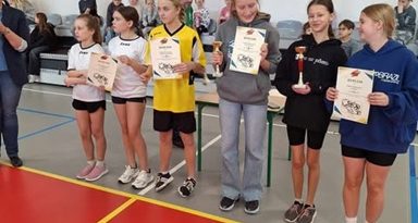 Zdjęcie przedstawia sześć zawodniczek z pięciu szkół, które odebrały dyplomy i puchary z zdobyte miejsca w Gminnych Mistrzostwach Piłki Koszykowej klas 4-6. Zawodniczka SP Czerwonak ma ubraną szarą bluzę oraz długie spodnie. Trzyma w rękach dyplom oraz puchar za 3 miejsce. W tle zdjęcia widać trybuny na których siedzą kibice. Ściana jest biała a parkiet pomarańczowo-szary.