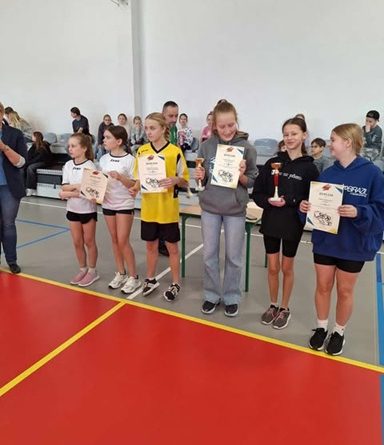 Zdjęcie przedstawia sześć zawodniczek z pięciu szkół, które odebrały dyplomy i puchary z zdobyte miejsca w Gminnych Mistrzostwach Piłki Koszykowej klas 4-6. Zawodniczka SP Czerwonak ma ubraną szarą bluzę oraz długie spodnie. Trzyma w rękach dyplom oraz puchar za 3 miejsce. W tle zdjęcia widać trybuny na których siedzą kibice. Ściana jest biała a parkiet pomarańczowo-szary.