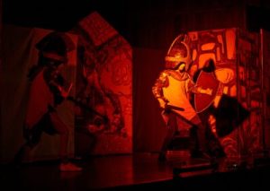 Scena pogrążona w półmroku ciepłego światła. Na scenie w dynamicznej pozycji stoją dwie postacie ubrane w kolczugi. W rękach trzymają miecze i tarcze.