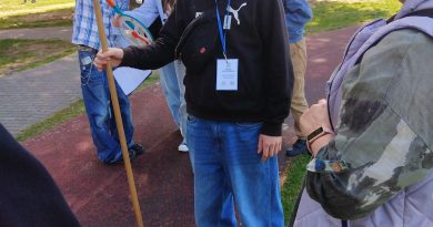 Na pierwszym planie widać chłopca stojącego z patykiem, który ma służyć do obliczenia wysokości. Z prawej strony stoi nauczyciel prowadzący warsztaty matematyczne w terenie. Z tyłu grupka uczniów odczytuje wykonany pomiar.