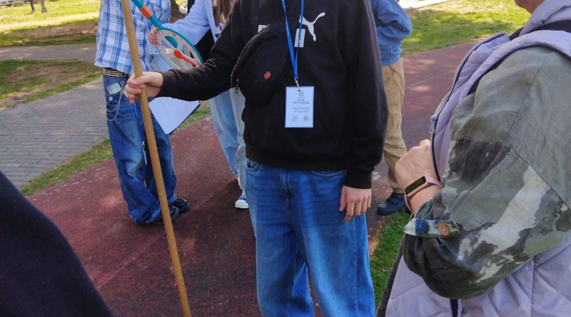 Na pierwszym planie widać chłopca stojącego z patykiem, który ma służyć do obliczenia wysokości. Z prawej strony stoi nauczyciel prowadzący warsztaty matematyczne w terenie. Z tyłu grupka uczniów odczytuje wykonany pomiar.