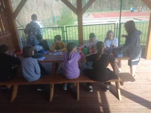 Grupa uczniów siedzących pod wiatą i wykonujących totemy z znalezionych skarbów przyrody. W oddali widać innych uczniów poszukujących skarbów.