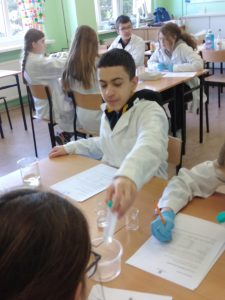 Na pierwszym planie widać chłopca w kitlu, który wykonuje doświadczenie chemiczne. Na stole stoją zlewki, leżą karty pracy. W oddali kolejna grupa uczniów dyskutuje na temat przeprowadzonego doświadczenia.