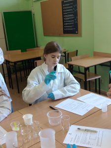 Na pierwszym planie widać uczennicę w kitlu, rękawiczkach, która potrząsając próbówką miesza jej zawartość.