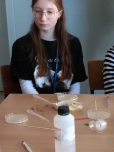 Na pierwszym planie uczennica słuchająca tłumaczenia nauczyciela o drożdżach. Na stole znajdują szalki Petriego, pipety, drożdże.