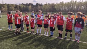 Uczniowie SP Czerwonak stojący w szeregu ubrani w czerwone koszulki z białymi rękawkami, czarne spodenki oraz piłkarskie getry odbierają medale z rąk organizatora zakładającego im medale na szyję.