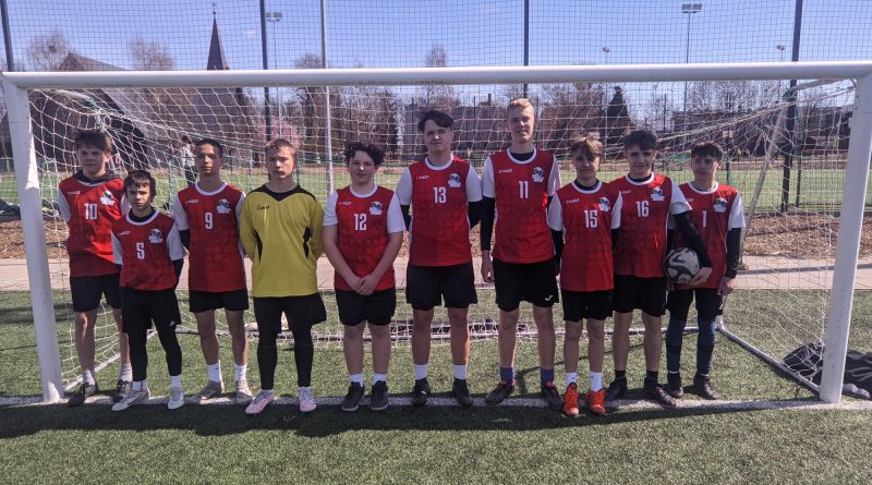 Na zdjęciu widzimy przedstawicieli drużyny SP Czerwonak ubranych w czerwone koszulki sportowe oraz czarne spodenki, ustawionych w szeregu przed bramką pozujących z piłką. Jako czwarty z lewej stoi bramkarz w żółtej bluzie.