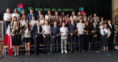 Absolwenci szkoły stoją w kilku rzędach na scenie. Trzymają róże