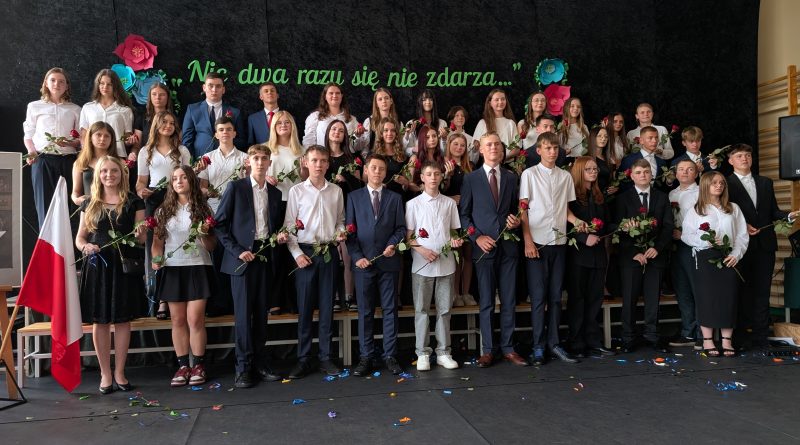 Absolwenci szkoły stoją w kilku rzędach na scenie. Trzymają róże