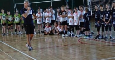 Przedstawia otwarcie turnieju. Trener UKS Czerwonak Karol Pabisiak otwiera II Turniej Minisiatkówki o Puchar Prezesa Klubu UKS Czerwonak. Zawodnicy są ustawieni na jasnym parkiecie. Ubrani są w swoje klubowe barwy. Reprezentanci UKS Czerwonak mają białe koszulki z czerwonymi dodatkami i czarne spodenki. W tle widać rozłożoną siatkę i boiska do kategorii „dwójki”. W głębi zdjęcia można dostrzec ciemnozieloną ścianę.