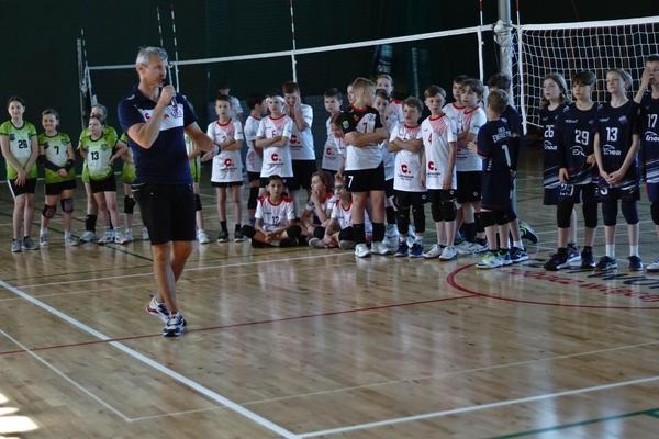 Przedstawia otwarcie turnieju. Trener UKS Czerwonak Karol Pabisiak otwiera II Turniej Minisiatkówki o Puchar Prezesa Klubu UKS Czerwonak. Zawodnicy są ustawieni na jasnym parkiecie. Ubrani są w swoje klubowe barwy. Reprezentanci UKS Czerwonak mają białe koszulki z czerwonymi dodatkami i czarne spodenki. W tle widać rozłożoną siatkę i boiska do kategorii „dwójki”. W głębi zdjęcia można dostrzec ciemnozieloną ścianę.