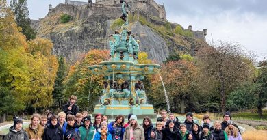 Grupa uczniów pozuje do wspólnego zdjęcia na schodach przy Pomniku Waltera Scotta w Edynburgu. W tle widać imponującą, gotycką wieżę i błękitne szkockie niebo.