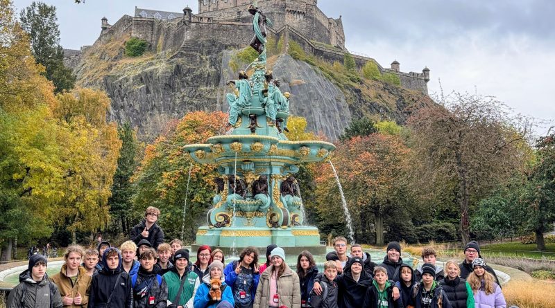 Grupa uczniów pozuje do wspólnego zdjęcia na schodach przy Pomniku Waltera Scotta w Edynburgu. W tle widać imponującą, gotycką wieżę i błękitne szkockie niebo.