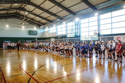 Przedstawia otwarcie zawodów. Widać na nim ustawione zawodniczki ubrane w kolorowe stroje sportowe. Przemawia Dyrektor Szkoły Podstawowej w Czerwonaku Pani Magdalena Zielenkiewicz. Dzieje się to na hali AKWEN w Czerwonaku. Wszyscy stoją na parkiecie a za plecami mają ustawioną siatkę oraz boiska do gry w minisiatkówkę.