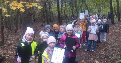 Uczniowie idący leśną ścieżką. Poszukują oznak jesieni, w ręku trzymają kartę pracy, na której zaznaczają odnalezione symbole jesieni. 