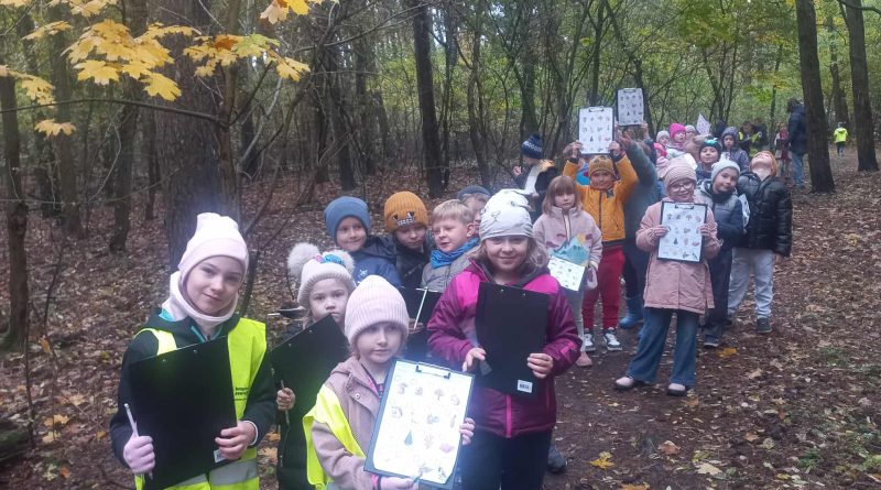 Uczniowie idący leśną ścieżką. Poszukują oznak jesieni, w ręku trzymają kartę pracy, na której zaznaczają odnalezione symbole jesieni. 