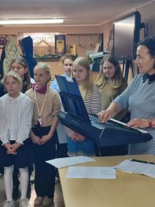 Na zdjęciu znajduje się grupa siedmiu odświętnie ubranych osób. Sześć dziewczynek i nauczycielka.

Od lewej strony stoi dziewczynka w białej bluzeczce i granatowej spódniczce. Tuż obok niej jest dziewczynka w beżowym sweterku i spodniach.

Po prawej stronie zdjęcia ustawiony jest instrument – keyboard. Na klawiszach gra nauczycielka ubrana w szarą sukienkę z czarnym, koronkowym kołnierzykiem. Obok nauczycielki stoi dziewczynka ma rozpuszczone włosy, ubrana jest w beżowy sweterek. Przed nią znajduje się blondynka w białym sweterku z zielonymi i czerwonymi paseczkami.

W drugim rzędzie stoją jeszcze dwie dziewczynki, uzupełniając całą grupę. Cała scena przedstawia przygotowania do wspólnego występu.