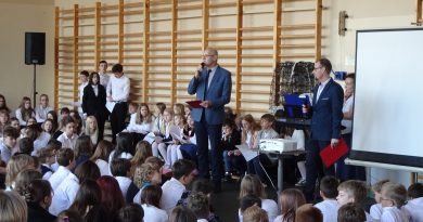 Zdjęcie przedstawia społeczność szkolną zebraną na sali gimnastycznej. Przed siedzącymi uczniami stoi dwóch nauczycieli historii przekazujących informacje dotyczące Narodowego Święta Niepodległości.