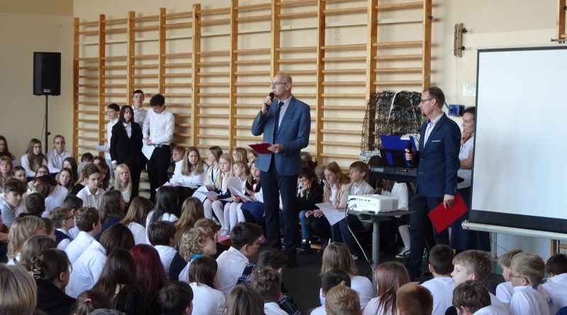 Zdjęcie przedstawia społeczność szkolną zebraną na sali gimnastycznej. Przed siedzącymi uczniami stoi dwóch nauczycieli historii przekazujących informacje dotyczące Narodowego Święta Niepodległości.