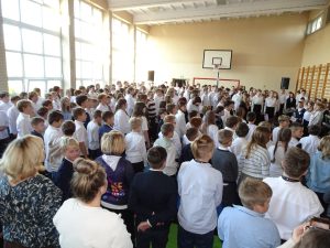 Zdjęcie przedstawia uczniów zgromadzonych w sali gimnastycznej w trakcie śpiewania hymnu państwowego. Dzieci stoją w postawie zasadniczej. Są odświętnie ubrani.