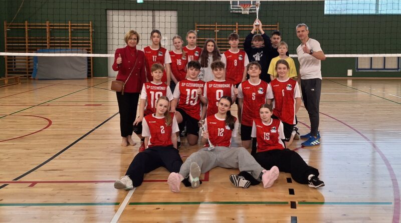 Zdjęcie to przedstawia zwycięską reprezentację dziewcząt i chłopców ze Szkoły Podstawowej w Czerwonaku. Są to uczniowie byłej klasy sportowej, która nie miała większych problemów aby wygrać w Gminnych Mistrzostwach Piłki Siatkowej klas 7-8. Ubrani są w stroje sportowe koloru czerwonego. Ustawieni w trzech szeregach. Pierwszy siedzi, drugi klęczy a trzeci stoi. Po lewej stronie stoi Pani Dyrektor Magdalena Zielenkiewicz a po prawej nauczyciel wychowania fizycznego Karol Pabisiak. W rękach uczniów znajdują się puchary za dwa pierwsze miejsca w zawodach. W tle widać siatkę oraz zieloną ścianę hali AKWEN w Czerwonaku.