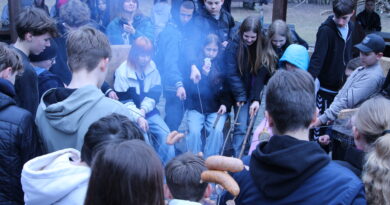 Uczniowie zgromadzeni wokół ogniska, w dłoniach trzymają kije i pieką kiełbaski. Widać unoszący się dym.