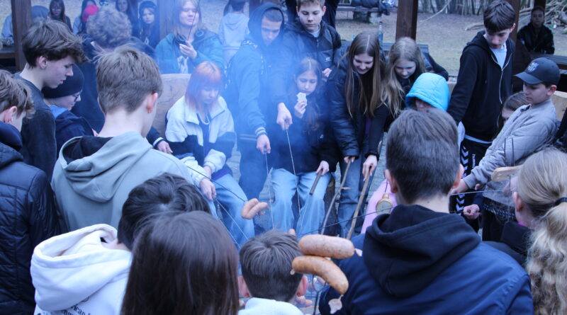Uczniowie zgromadzeni wokół ogniska, w dłoniach trzymają kije i pieką kiełbaski. Widać unoszący się dym.