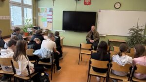 prowadzący siedzi pod tablicą zwrócony twarzą do klasy