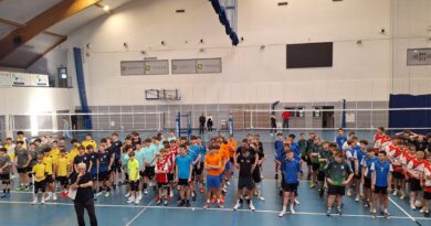 To zdjęcie grupowe chłopaków. 17 reprezentacji z całego powiatu poznańskiego stoi przed siatką do wspólnego zdjęcia. Wszyscy zawodnicy mają na sobie wielokolorowe stroje sportowe w barwach swoich szkół. Na centralnym miejscu zdjęcia widać uczniów SP Czerwonak. Posiadają biało-czerwony ubiór. Parkiet hali w Rokietnicy jest granatowo-szary. Ściany jasna.