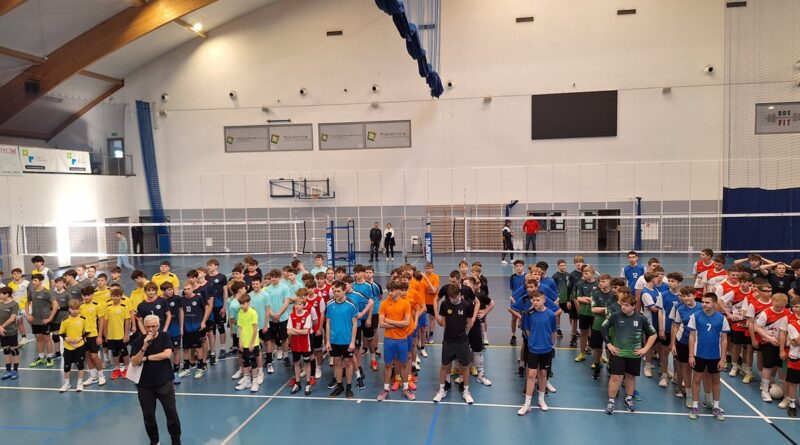 To zdjęcie grupowe chłopaków. 17 reprezentacji z całego powiatu poznańskiego stoi przed siatką do wspólnego zdjęcia. Wszyscy zawodnicy mają na sobie wielokolorowe stroje sportowe w barwach swoich szkół. Na centralnym miejscu zdjęcia widać uczniów SP Czerwonak. Posiadają biało-czerwony ubiór. Parkiet hali w Rokietnicy jest granatowo-szary. Ściany jasna.