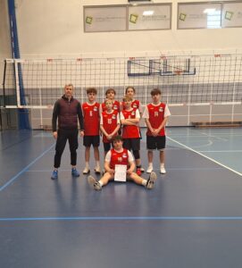 To zdjęcie przedstawiam nauczyciela oraz trenera piłki siatkowej, który jest ustawiony do wspólnego zdjęcia wraz ze swoimi podopiecznymi z klasy 8a. Sześciu zawodników stoi i jeden siedzi z dyplomem. Ubrani są w strój sportowy w barwach SP Czerwonak. Biało czerwonym. Trener ma na sobie czarny ubiór i brązową kamizelkę. W tle widać siatkę oraz jasną ścianę hali w Rokietnicy.