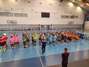 Zdjęcie 5.

To zdjęcie grupowe z turnieju siatkówki chłopców Igrzyszk Dzieci na szczeblu powiatowym. Wszystkie reprezentacje ustawione są w rzędach i mają na sobie stroje sportowe w barwach swoich szkół. Zawodnicy i zarazem uczniowie z klasy sportowej 5c z SP Czerwonak mają czerwono białe stroje. Widać również organizatora zawodów Pana Piotra Paupa z Rokietnicy. W tle jest rozłożona siatka i boiska przygotowane do rozgrywek. Parkiet jest koloru granatowo-szarego. Ściany jasne.