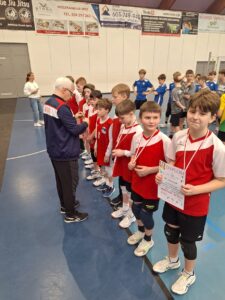 To zdjęcie przedstawia wręczenie medali oraz dyplomu reprezentacji szkoły podstawowej z Czerwonaka. Zawodnicy ustawieni są w szeregu i przyjmują gratulacje i medale od organizatora zawodów Pana Piotra Paupa. Ubrani są w strój sportowy biało czerwony. Za nimi znajdują się reprezentacje z miejsca drugiego i pierwszego.