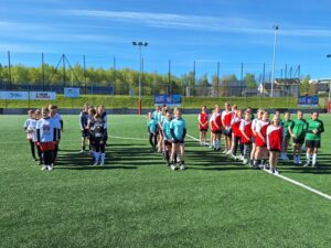 Na tym zdjęciu widać zawodniczki z pięciu szkół, które biorą udział w turnieju. Stoją w rzędzie i ubrane są w stroje sportowe zaczynając od lewej: białe, czarne, błękitne, biało-czerwone, zielone. Stoją na płycie boiska w kolorze zielonym. U góry widać błękitne, słoneczne niebo.