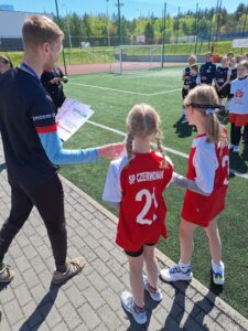 Na tym zdjęciu dwie dziewczyny z SP Czerwonak odbierają dyplom od organizatora zawodów AKWEN reprezentowanego przez Pana Wojciecha Grabińskiego. Pan Wojciech ubrany jest w czarny strój sportowy. Trzyma dyplomy, które wręcza za poszczególne miejsca. Dziewczyny z SP Czerwonak są w strojach czerwono-białych. W tle widać inne zawodniczki, które stoją na zielonej, sztucznej murawie stadionu Orlik w Czerwonaku.