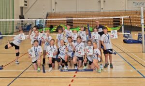 Na zdjęciu widać wszystkich zawodników i zawodniczki biorących udział w XVI edycji ENEA Mini CUP. Jest to zdjęcie zbiorowe. Wszyscy mają na sobie białe koszulki otrzymane od organizatora ENEA ENERGETYK Poznań. Ustawieni są pod siatką. W tle widać kolorowy, duży tor przeszkód „dmuchaniec” z którego korzystały dzieci podczas zawodów.