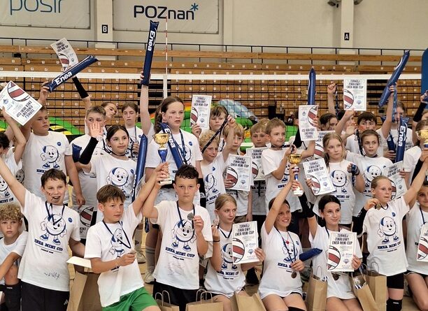 Na zdjęciu widać wszystkich zawodników i zawodniczki biorących udział w XVI edycji ENEA Mini CUP. Jest to zdjęcie zbiorowe. Wszyscy mają na sobie białe koszulki otrzymane od organizatora ENEA ENERGETYK Poznań. Ustawieni są pod siatką. W tle widać kolorowy, duży tor przeszkód „dmuchaniec” z którego korzystały dzieci podczas zawodów.