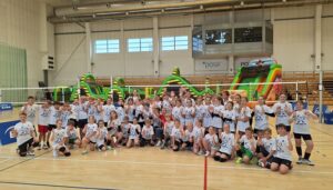Zdjęcie przedstawia uczniów wszystkich szkół biorących udział w XVI edycji ENEA Mini CUP. Pierwszy szereg jest w pozycji klęczącej. Drugi stoi. Wszyscy mają białe koszulki organizatora z logo ENEA Mini CUP. Zawodnicy i zawodniczki trzymają w rękach liczne nagrody oraz puchary. W tle widać rozwieszoną siatkę do gry sportowej piłka siatkowa.